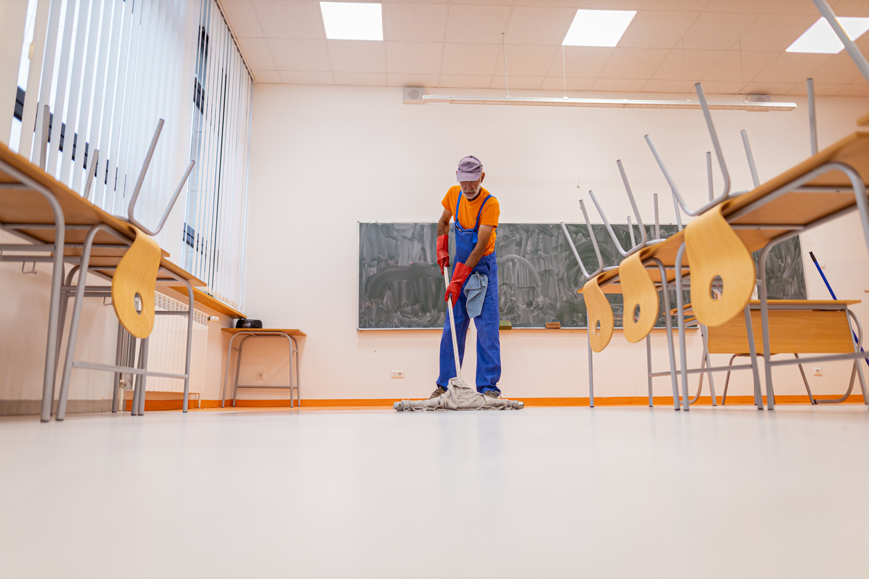 A front view of an unrecognizable mature janitor mopping the floor of an empty education classroom.